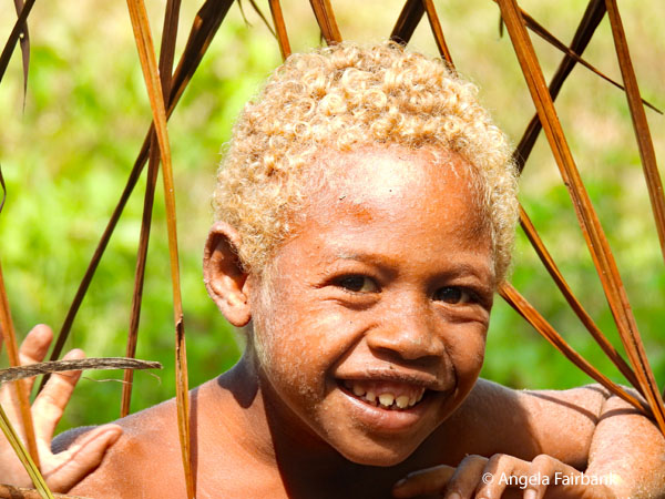 grinning blong-haired boy behind grass fence 